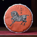 A round cake of Pu Er tea wrapped in an orange paper sleeve featuring a black horse illustration and Chinese characters, displayed on a wooden table with a teapot and ceramic vessel in a dark, traditional setting.