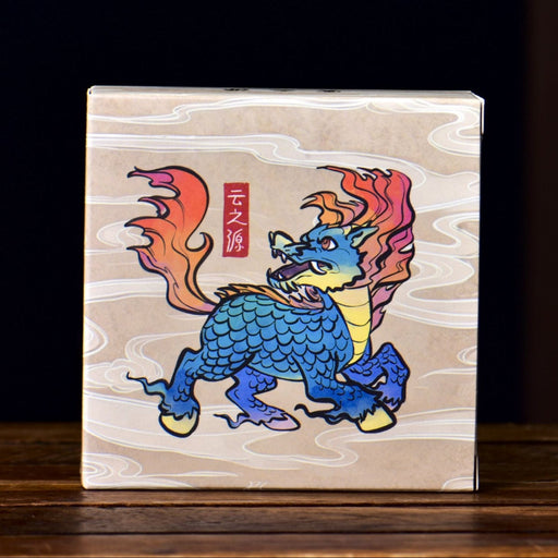 A square box containing ripe pu-erh tea with a light wood-grain background featuring a colorful illustration of a blue dragon with orange and red flames, standing on a dark wooden surface.