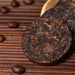 Dark, compressed tea cakes with visible leaf texture arranged in a wooden scoop on a brown wooden surface, with scattered coffee beans around them.
